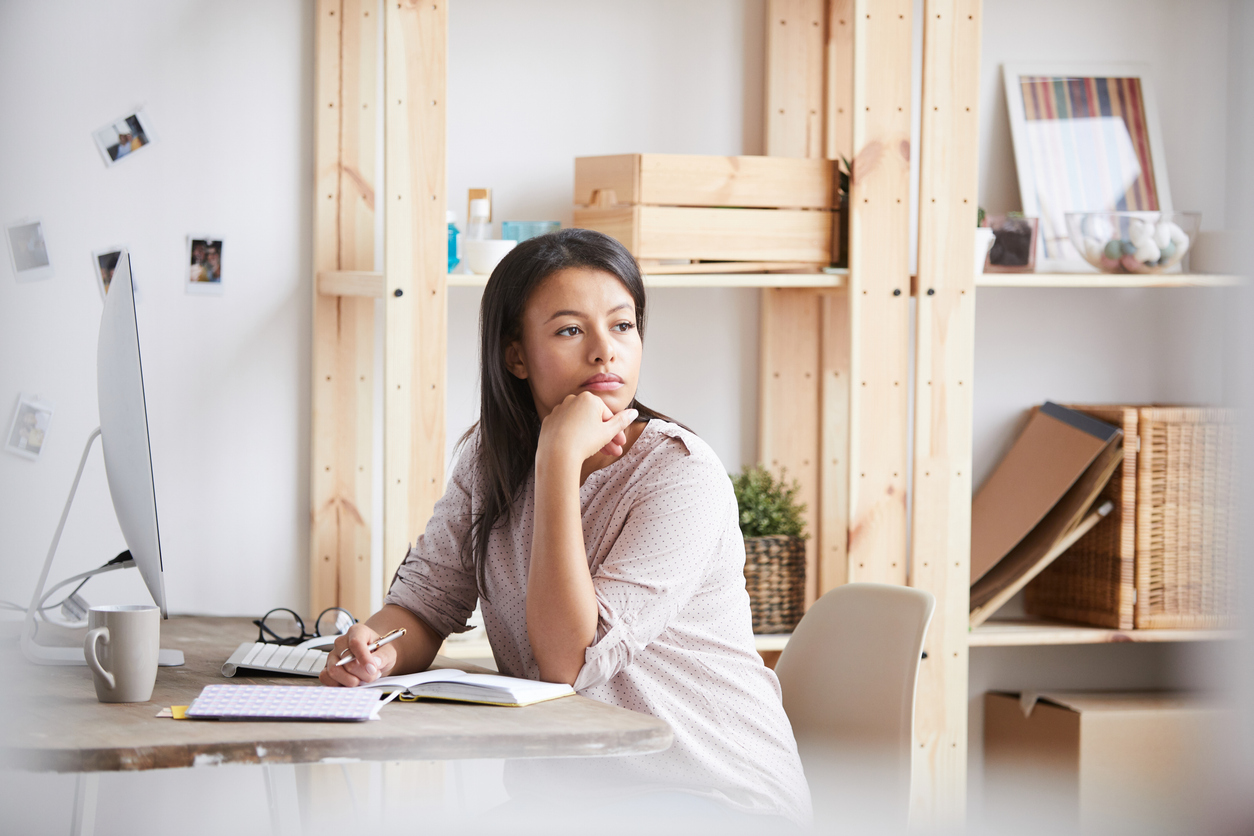 Woman sitting at her desk with a notebook, looking thoughtful and reflective while working from home.