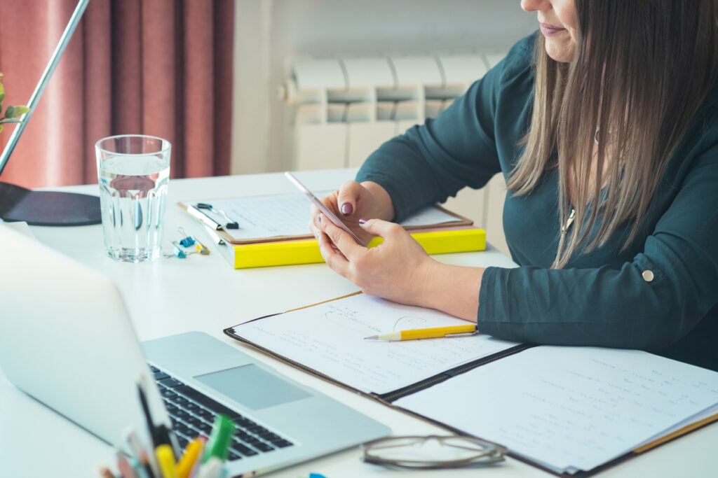 Person sitting at a desk using their phone while work remains unfinished on a laptop and papers, illustrating productive procrastination